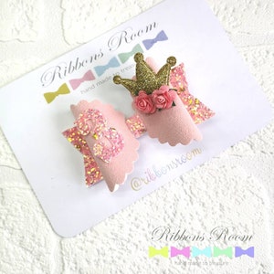 K&ouml;nnte beinhalten: Zwei rosa Haarschleifen mit Glitzerakzenten. Eine Schleife hat eine goldene Glitzerkrone mit rosa Rosen, die andere eine Glitzer-8. Die Schleifen befinden sich auf einer wei&szlig;en Karte mit dem Text "Ribbons Room".