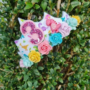 May include: A pink glittery number three on a white crown with a dinosaur print. The crown is decorated with colorful flowers.