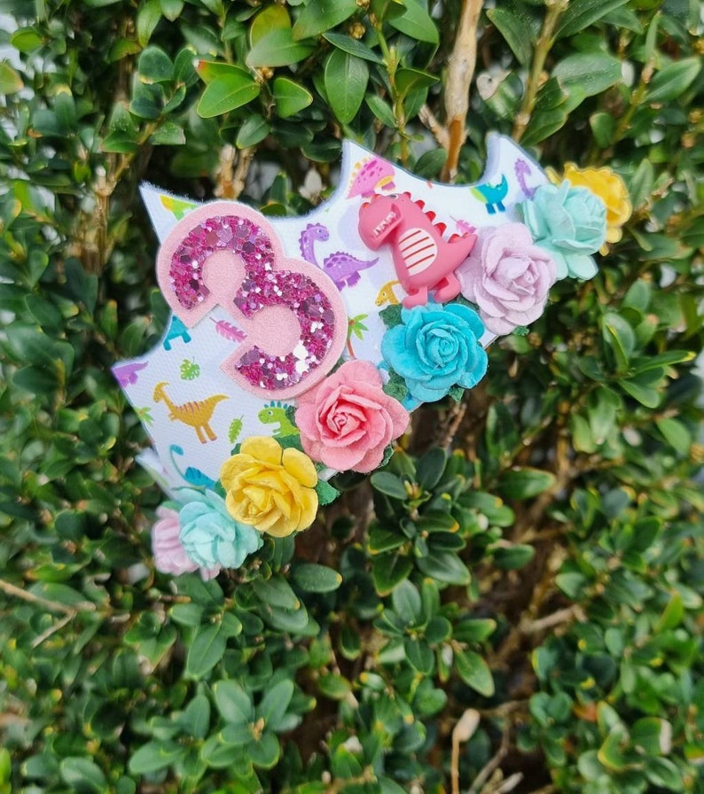 May include: A pink glittery number three on a white crown with a dinosaur print. The crown is decorated with colorful flowers.