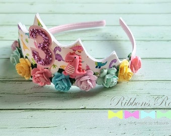 May include: A white headband with a pink, green, and yellow flower crown. The crown features a pink number three with glitter and a pink dinosaur. The headband says "Ribbons Room" and "Hand made to treasure".