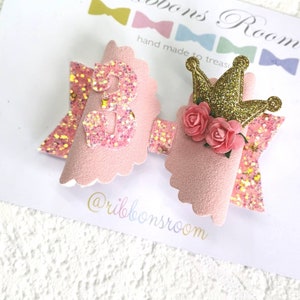 K&ouml;nnte beinhalten: Eine rosa Haarschleife mit einer glitzernden Zahl 3, einer goldglitzernden Krone und rosa Rosen. Die Schleife hat einen gewellten Rand und einen glitzernden Akzent. Der Text "Ribbons Room" ist sichtbar.