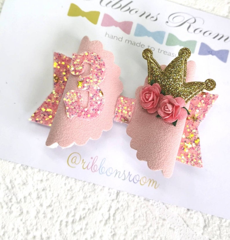 K&ouml;nnte beinhalten: Eine rosa Haarschleife mit einer glitzernden Zahl 3, einer goldglitzernden Krone und rosa Rosen. Die Schleife hat einen gewellten Rand und einen glitzernden Akzent. Der Text "Ribbons Room" ist sichtbar.