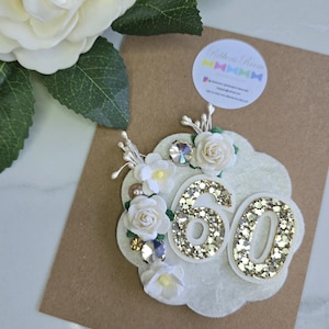 May include: A decorative felt badge featuring the number 60 in gold glitter, accented with small white fabric roses, green leaves, clear crystals, and pearl-like beads. The scalloped white felt base is mounted on a brown card with a circular logo sticker.