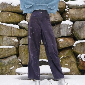 Könnte beinhalten: Eine Frau trägt einen hellblauen, kurzen Pullover mit zwei Taschen und eine dunkelgraue, weite Hose. Sie steht vor einer mit Schnee bedeckten Steinmauer. Der Text "DongoDesign" ist in der unteren rechten Ecke des Bildes sichtbar.