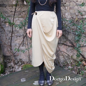 Può includere: Una donna indossa una camicia nera a maniche lunghe con una collana d'oro e una gonna avvolgente beige. La gonna ha una grande tasca sul davanti ed è legata in vita con una cintura marrone. È in piedi su un ponte di legno di fronte a un muro di mattoni con dei rampicanti.