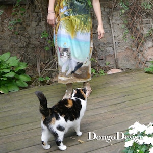 May include: A woman wearing a blue and green maxi dress with a palm tree and cat print stands on a wooden deck. A calico cat sits in front of her, looking up at her.