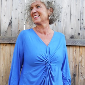 Könnte beinhalten: Ein leuchtend blaues Langarm-Oberteil mit V-Ausschnitt und einem geknoteten Detail an der Taille. Das Oberteil wird mit einer blauen Jeans kombiniert. Der Text "DongoDesign" ist unten rechts sichtbar.