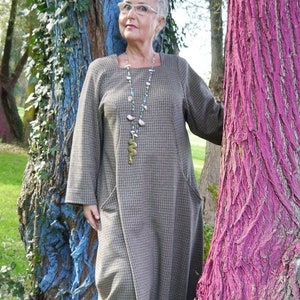 May include: A woman wearing a brown and white checkered dress with long sleeves and a square neckline. She is standing in front of two trees, one painted blue and the other pink. She is wearing a long necklace with a snake pendant and glasses. The text "DongoDesign" is visible in the bottom right corner of the image.