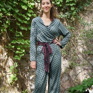 Könnte beinhalten: Eine Frau trägt einen grünen und schwarzen Polka-Dot-Wickel-Jumpsuit mit einem burgunderfarbenen und schwarzen gemusterten Gürtel. Sie steht auf einem Holzdeck mit grünem Laub im Hintergrund.