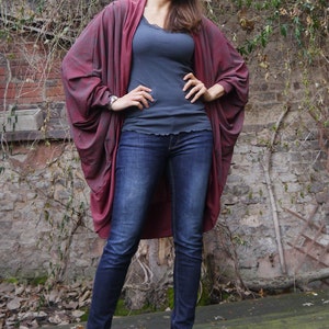 May include: A woman wearing a long, burgundy-colored cardigan with a gray tank top and blue jeans. She is standing in front of a brick wall with vines growing on it. The text "DongoDesign" is visible in the bottom right corner of the image.