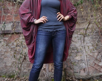 Könnte beinhalten: Eine Frau trägt einen langen, burgunderfarbenen Cardigan mit einem grauen Tanktop und blauen Jeans. Sie steht vor einer Backsteinmauer mit Ranken, die daran wachsen. Der Text "DongoDesign" ist in der rechten unteren Ecke des Bildes sichtbar.