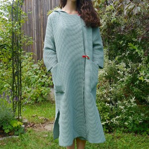 Op de afbeelding: Een lange, gekapte robe in een lichtgroen en wit geruit patroon. De robe heeft een enkele knoopsluiting en twee zakken. Het model staat in een tuinomgeving.