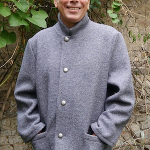 Puede incluir: Una chaqueta de lana gris con cuello alto y cinco botones plateados. La chaqueta tiene dos bolsillos de parche y un logotipo de DongoDesign en la parte inferior derecha.