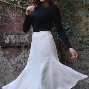 May include: A woman wearing a black turtleneck sweater and a white, flowing skirt. The skirt has a slight flare at the bottom. The woman is smiling and looking at the camera. The image is taken outdoors in front of a brick wall.