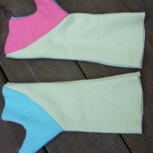 May include: Two pairs of knitted arm warmers with contrasting colored panels. One pair is cream with a pink panel and the other is cream with a blue panel. The arm warmers are laid out on a wooden surface.