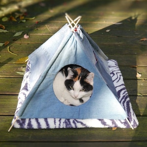 Puede incluir: Un gato está descansando dentro de una tienda de campaña tipi de mezclilla azul con una base a rayas blancas y negras. El tipi está en una plataforma de madera.