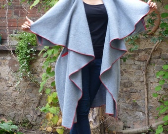 May include: A gray wrap coat with a red trim. The coat is draped over the shoulders and has a long, flowing design. The model is wearing blue jeans and white heels.