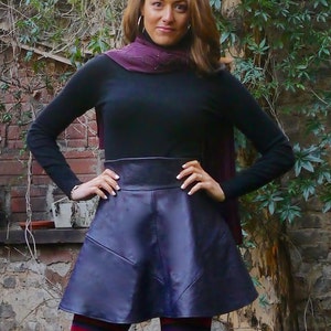 May include: A woman wearing a black turtleneck sweater with a purple scarf, a dark purple leather skirt, and colorful striped tights. The skirt has a high waist and a flared silhouette. The woman is standing in front of a brick wall with greenery.