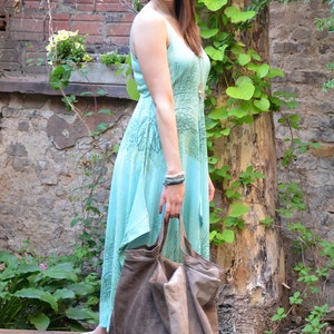 May include: A woman wearing a light blue maxi dress with a leafy print and a brown tote bag. The dress has a loose, flowing fit and a slightly asymmetrical hemline. The woman is standing in front of a brick wall with green vines growing up it.