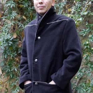 May include: A black wool jacket with a white trim and a high collar. The jacket has a button closure and two pockets. The jacket is being worn by a person standing in front of a green leafy background.