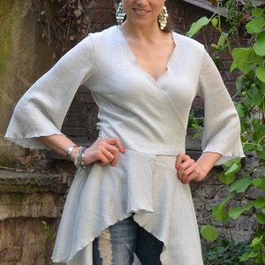 Pode incluir: Um top prateado de amarrar com uma bainha franzada e mangas compridas. O top é assimétrico e tem um decote em V. A modelo está usando o top com jeans desfiados.
