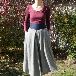 May include: A woman wearing a burgundy long-sleeved shirt with a lace neckline and a gray skirt with a black waistband. She is standing in front of a green bush and a white building.