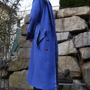 May include: A long blue coat with a hood, yellow trim, and brown buttons. The coat is worn by a person standing in front of a stone wall.