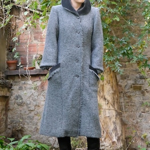 May include: A long gray wool coat with a black collar and black trim on the pockets. The coat has a button closure and is worn by a person with black boots.