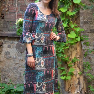 Puede incluir: Una mujer lleva un vestido midi estampado de colores con mangas acampanadas. El vestido tiene una variedad de colores, incluyendo azul, rojo y negro. La mujer está de pie frente a una pared de ladrillo con enredaderas verdes.