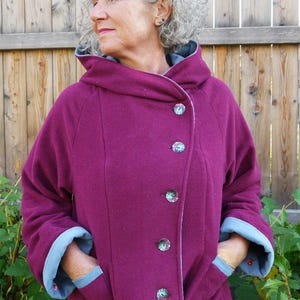 May include: A plum-colored hooded jacket with a wrap-style front, featuring decorative buttons and light blue trim on the hood and cuffs. The jacket has pockets and is made of a soft, textured fabric. The brand name "Dongo Design" is visible.