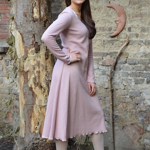 Könnte beinhalten: Ein rosafarbenes, langärmeliges Kleid mit V-Ausschnitt und einem gewellten Saum. Das Kleid ist aus einem weichen, bequemen Stoff gefertigt und hat eine lockere Passform. Das Model trägt weiße Strumpfhosen und rosa Schuhe.