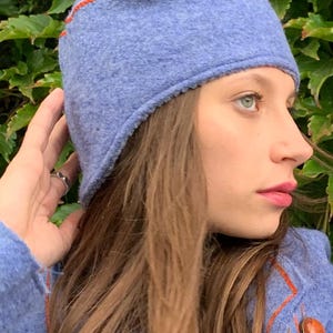 May include: A blue wool hat with a red trim. The hat is worn by a person with long brown hair. The hat has a flap that covers the ears. The hat is made by DongoDesign.