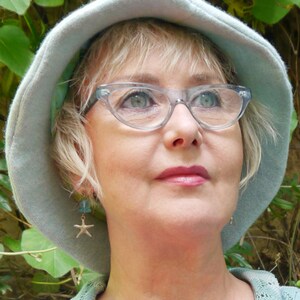May include: A woman wearing a light blue hat with a wide brim, clear cat-eye glasses, and a light blue and white patterned sweater. She is wearing star-shaped earrings.