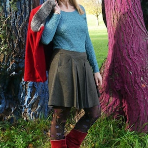 May include: A woman wearing a teal sweater, a gray skirt, red boots, and patterned tights stands between two trees. The trees are painted blue and pink.