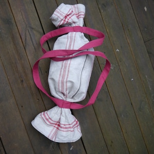 Puede incluir: Una bolsa de tela blanca con rayas rojas y un cierre de cordón rosa. La bolsa tiene forma de caramelo y está sobre una superficie de madera.
