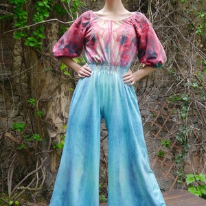 Könnte beinhalten: Eine Frau trägt einen blau-grünen Jumpsuit mit einem Oberteil mit Blumendruck. Das Oberteil hat einen runden Ausschnitt und Puffärmel. Der Jumpsuit hat eine weite Hose und eine taillierte Taille.