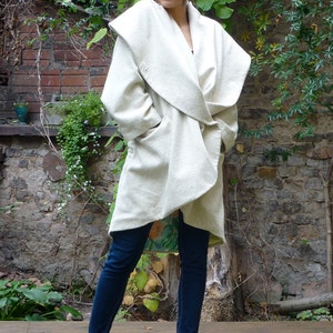 May include: A white wrap coat with a large collar and a tie belt. The coat is made of a soft, textured fabric. The model is wearing blue jeans and beige shoes.