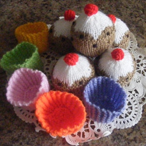 Puede incluir: Cupcakes tejidos a mano con glaseado blanco, bizcocho marrón y una cereza roja encima. Los cupcakes están rodeados de coloridos moldes de cupcakes tejidos en naranja, rosa, verde, amarillo y azul. Dispuestos sobre un mantel individual de encaje blanco.