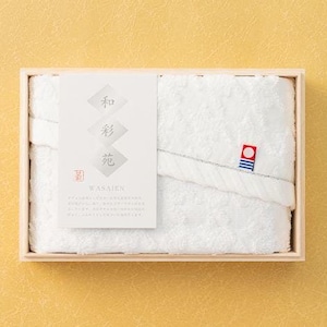 May include: A white towel set neatly arranged in a wooden box. The towels are folded and feature a decorative label with Japanese text and a small emblem. The box is presented on a textured yellow background.