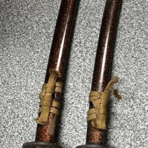 May include: Two Japanese-style swords with brown and gold accents. The blades have a dark, speckled finish. The handles are wrapped in a gold-colored material. The swords are of different sizes.