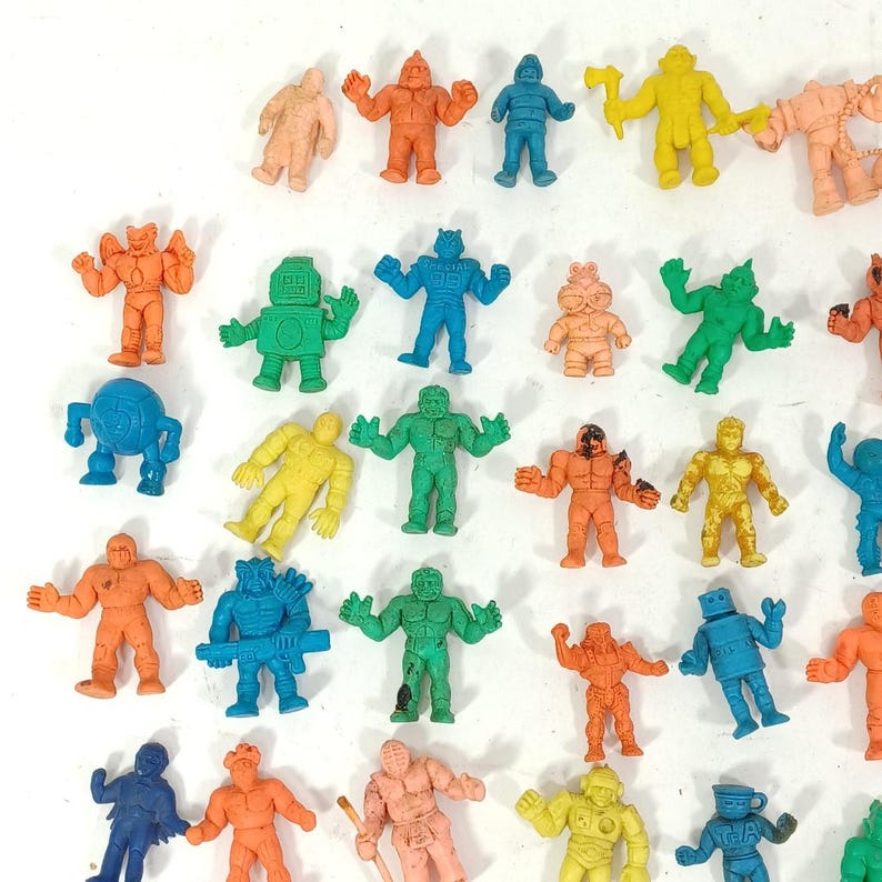 May include: A collection of small, colorful action figure toys in various poses. The figures are in shades of orange, blue, green, yellow, and tan. Some figures have robot-like designs, while others resemble muscular characters. The toys are made of a rubbery material.