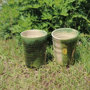 May include: Two green and white ceramic mugs with a speckled glaze. The mugs are sitting on a bed of green grass.
