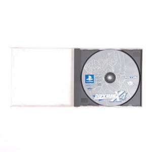 Puede incluir: Un disco de juego de PlayStation plateado con una ilustraci&oacute;n en blanco y negro de personajes del videojuego Rockman X4. El disco tiene el texto "Rockman X4" y "CAPCOM" impreso en &eacute;l.