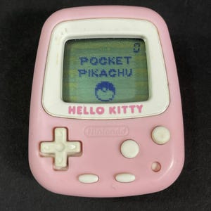 May include: A pink handheld electronic game device featuring a digital screen displaying the text POCKET PIKACHU and a Poké Ball icon. The device has the text HELLO KITTY printed below the screen, a Nintendo logo, a directional pad, and buttons.