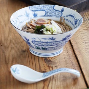 以下が含まれることがあります： 青い波と花の模様が入った白いセラミックボウル。麺、肉、野菜が入っています。青い花の模様が入ったお揃いのスプーンがボウルの横に置かれています。木製の表面にあります。