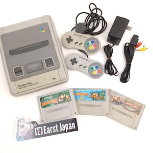 Może przedstawiać: Szara konsola Nintendo Super Famicom z dwoma pasującymi kontrolerami, zasilaczem i kartridżami do gier. Kontrolery mają kolorowe przyciski i pady kierunkowe. Kartridże zawierają tytuły gier i kolorową grafikę.