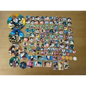 May include: A collection of Dragon Ball Z collectible items, including circular game pieces and trading cards. The pieces feature colorful illustrations of characters from the anime series. The items are arranged on a wooden surface.
