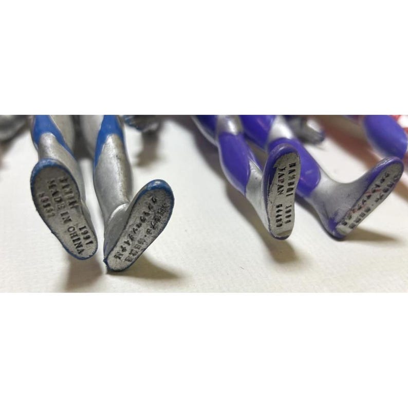 May include: Two pairs of silver and blue plastic action figure legs. The left pair has text on the soles reading 1991 MADE IN CHINA. The right pair has text on the soles reading BANDAI 1991 JAPAN 64489, with additional Japanese characters.