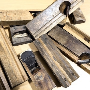 May include: A collection of vintage wooden hand planes, some with metal blades, used for woodworking. The planes are in various sizes and shapes, with a worn and weathered appearance.
