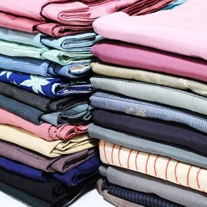 Op de afbeelding: Een stapel gevouwen stof in verschillende kleuren en patronen, waaronder roze, blauw, groen en beige. De stof is netjes gestapeld, met de randen van elk stuk zichtbaar.
